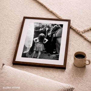 May include: A framed black and white photograph of a young girl standing in front of a group of men. The frame is dark brown, and a beige mug of coffee sits nearby. A beige pillow is in the foreground.