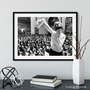 Puede incluir: Impresi&oacute;n enmarcada en blanco y negro de una mujer sonriendo y agitando una prenda sobre una multitud. La fotograf&iacute;a vintage es probablemente de mediados del siglo XX. La impresi&oacute;n se exhibe sobre una superficie negra con objetos decorativos.