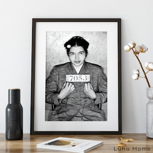 May include: A black and white framed portrait of Rosa Parks wearing glasses and a collared jacket, holding a sign with the number 7053. The print is displayed in a dark frame, capturing a historical mugshot style image with clear details.