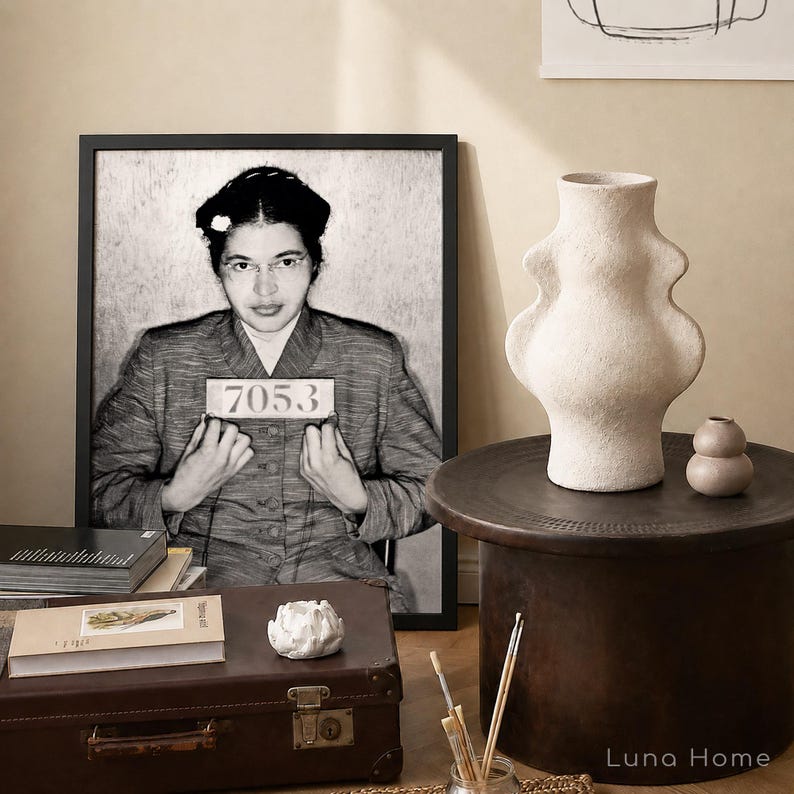 May include: A framed black and white portrait of Rosa Parks holding a booking sign with the number 7053. Next to it is a textured off-white ceramic vase with a curved silhouette, a small round beige vase, a brown suitcase, and books.