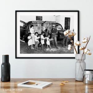 May include: Black and white photograph of a mobile library. A group of children and adults are gathered around a car with open doors, filled with books. The children are holding books and reading.