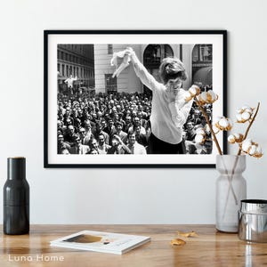 Puede incluir: Impresi&oacute;n enmarcada en blanco y negro que muestra a una mujer sonriendo y sosteniendo una prenda, con una gran multitud al fondo. La fotograf&iacute;a parece ser una imagen vintage, posiblemente tomada en una ciudad.