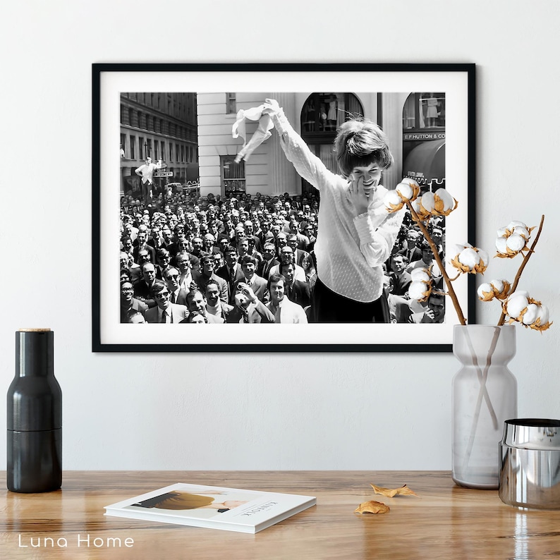 Puede incluir: Impresi&oacute;n enmarcada en blanco y negro que muestra a una mujer sonriendo y sosteniendo una prenda, con una gran multitud al fondo. La fotograf&iacute;a parece ser una imagen vintage, posiblemente tomada en una ciudad.