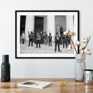 May include: Black and white framed photograph of a group of people standing on the steps of a building. Some individuals are holding rifles. The photograph is displayed on a wooden surface with decorative items.