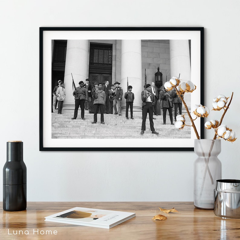 May include: Black and white framed photograph of a group of people standing on the steps of a building. Some individuals are holding rifles. The photograph is displayed on a wooden surface with decorative items.