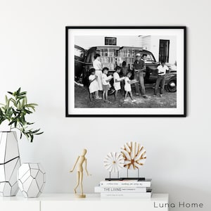 May include: Black and white framed photograph depicting a group of children and adults gathered around a vehicle filled with books. The image captures a moment of community and education, with people holding books.