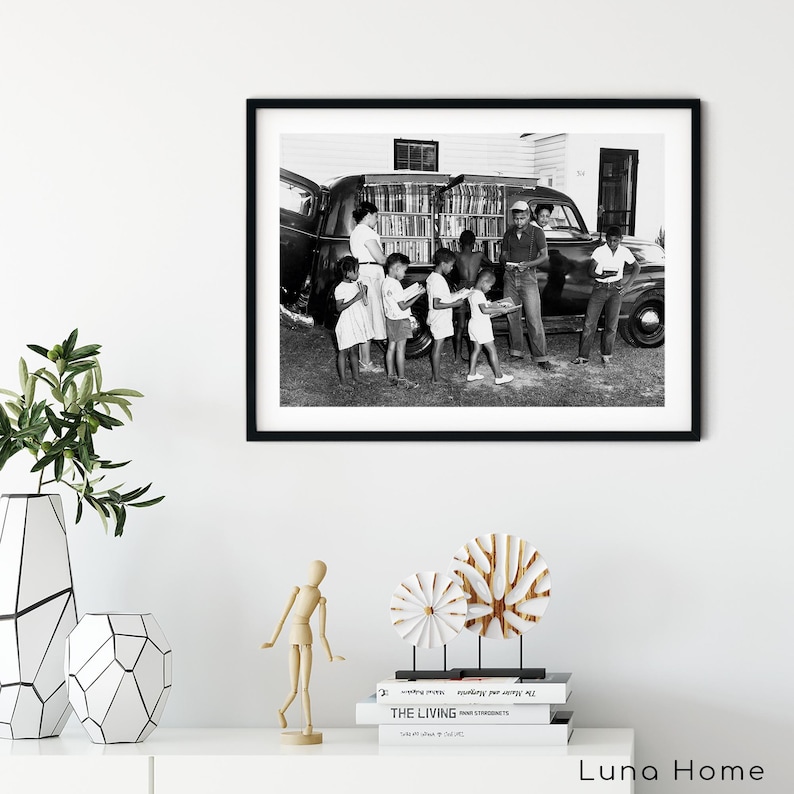 May include: Black and white framed photograph depicting a group of children and adults gathered around a vehicle filled with books. The image captures a moment of community and education, with people holding books.