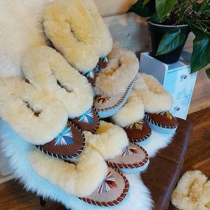 Könnte beinhalten: Eine Sammlung gemütlicher Schaffell-Hausschuhe in verschiedenen Größen. Die Hausschuhe haben eine hellbraune Wildlederbasis mit farbenfrohen Stickereien und ein flauschiges, cremefarbenes Schaffellfutter. Sie sind auf einer weißen Kunstfelloberfläche angeordnet.