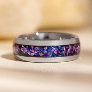May include: A grey tungsten carbide ring with a band of purple and blue glitter embedded in black resin. The ring is on a light beige surface. "REDWOOD RINGS" is printed in black below the ring.