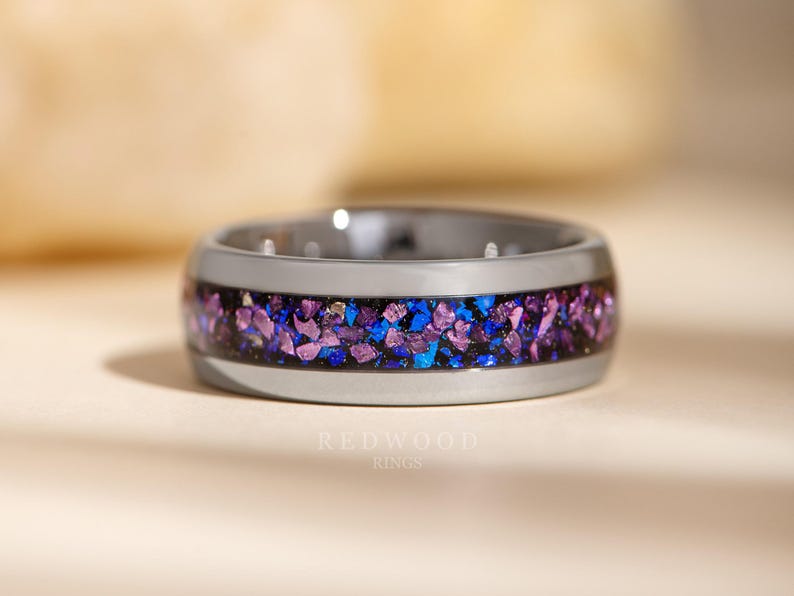 May include: A grey tungsten carbide ring with a band of purple and blue glitter embedded in black resin. The ring is on a light beige surface. "REDWOOD RINGS" is printed in black below the ring.