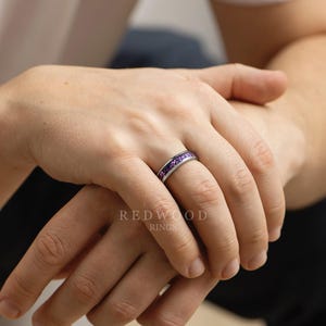 May include: A silver ring with a band of small, purple gemstones. The ring is on a person's finger, with the Redwood Rings logo visible in the background. The ring is a piece of jewelry.