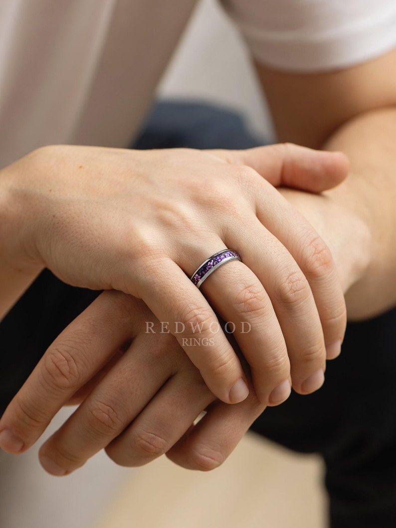 May include: A silver ring with a band of small, purple gemstones. The ring is on a person's finger, with the Redwood Rings logo visible in the background. The ring is a piece of jewelry.