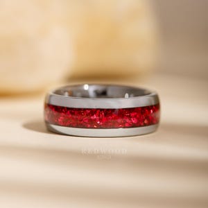 May include: A silver-colored ring with a wide band. The center of the ring is filled with a vibrant red, textured material. The ring is a close-up shot, with the brand name "REDWOOD RINGS" visible at the bottom.