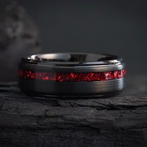 Peut inclure: Gros plan sur une bague noire ornée d'une bande de pierres rouge vif. La bague est mise en valeur sur un fond sombre et texturé, soulignant le contraste entre le métal poli et les pierres précieuses éclatantes. Design moderne et audacieux.