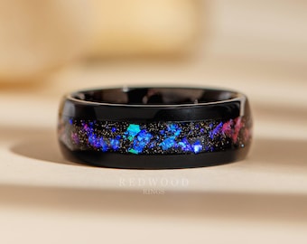 May include: A black ceramic ring with a thin band of iridescent blue, pink, and gold glitter embedded in the black ceramic. The ring is on a light beige surface.  REDWOOD RINGS