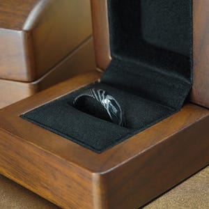 May include: A Damascus steel ring with a unique pattern rests in a wooden ring box with a black velvet interior. The box is open, revealing the ring's intricate design. The box is made of dark wood.