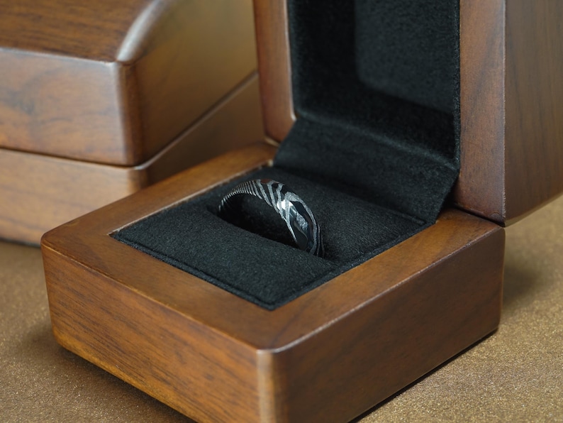 May include: A Damascus steel ring with a unique pattern rests in a wooden ring box with a black velvet interior. The box is open, revealing the ring's intricate design. The box is made of dark wood.