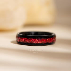 May include: A black ring with a band of vibrant red stones. The ring has a polished finish and the text "REDWOOD RINGS" is visible on the surface.