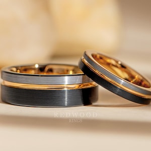 Puede incluir: Dos alianzas de carburo de tungsteno con acabado cepillado. Una alianza tiene una incrustación dorada y la otra una incrustación plateada. Las alianzas están sobre una superficie blanca.