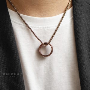 May include: A bronze ring pendant necklace. The circular pendant hangs from a matching chain. The necklace is worn over a white t-shirt and a dark jacket. The brand name "REDWOOD RINGS" is visible in the lower left corner.