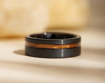 May include: A close-up of a black ring with a copper-coloured wood inlay. The ring has a brushed metal finish and the text "REDWOOD RINGS" is visible at the bottom.