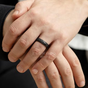 May include: A black ring with a textured pattern is displayed on a person's finger. The ring has a dark, matte finish and a subtle, organic design. The brand name "REDWOOD RINGS" is visible in the image.