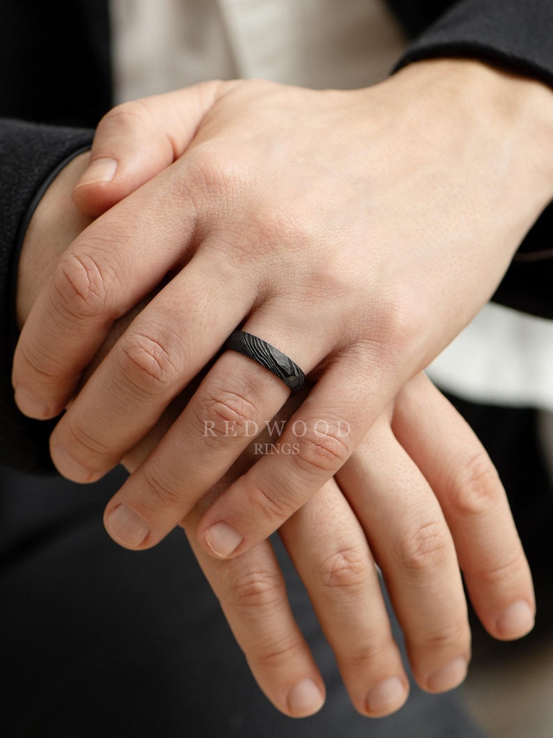 May include: A black ring with a textured pattern is displayed on a person's finger. The ring has a dark, matte finish and a subtle, organic design. The brand name "REDWOOD RINGS" is visible in the image.