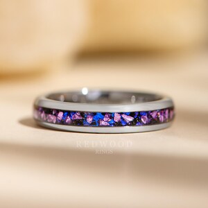 May include: A silver ring with a band of purple and blue glitter embedded in the metal. The ring is on a light brown surface.