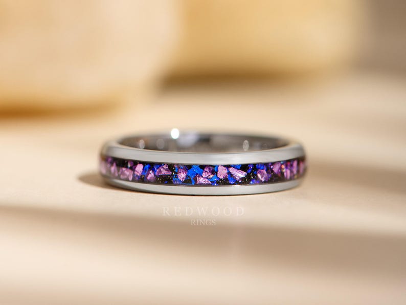 May include: A silver ring with a band of purple and blue glitter embedded in the metal. The ring is on a light brown surface.