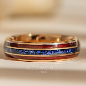 May include: A gold-colored ring with a band of alternating red and blue inlays. The blue inlay appears to be made of crushed stone. The ring is a close-up shot, with the brand name "REDWOOD RINGS" visible at the bottom.
