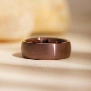 May include: A close-up of a brushed copper ring. The ring has a wide band and a smooth, matte finish. The text "REDWOOD RINGS" is visible at the bottom of the image.