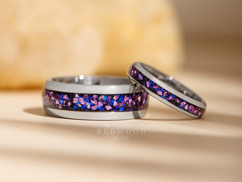 May include: Two silver tungsten carbide rings with a black line and a band of purple and blue glitter. The rings are on a light brown surface.