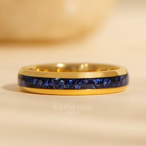 May include: A gold-colored ring with a band of deep blue, textured inlay. The ring has a polished finish and is set against a soft, neutral background. The text "REDWOOD RINGS" is visible.