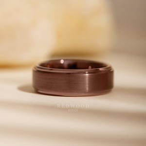 May include: A close-up of a rose gold ring with a brushed metal finish. The ring has a flat top and a slightly raised outer edge. The brand name "REDWOOD RINGS" is printed in white at the bottom.