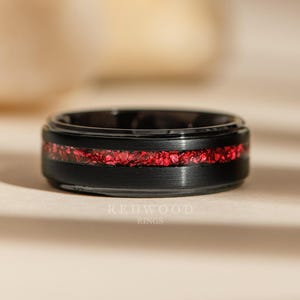 Puede incluir: Un anillo negro con una incrustación roja vibrante. El anillo tiene un acabado de metal cepillado y una banda de piedras o cristales rojos. El logotipo de Redwood Rings es visible en la parte inferior.