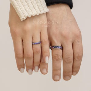 May include: Two matching rings with a band of small purple and blue gemstones. The rings are on the fingers of two people.