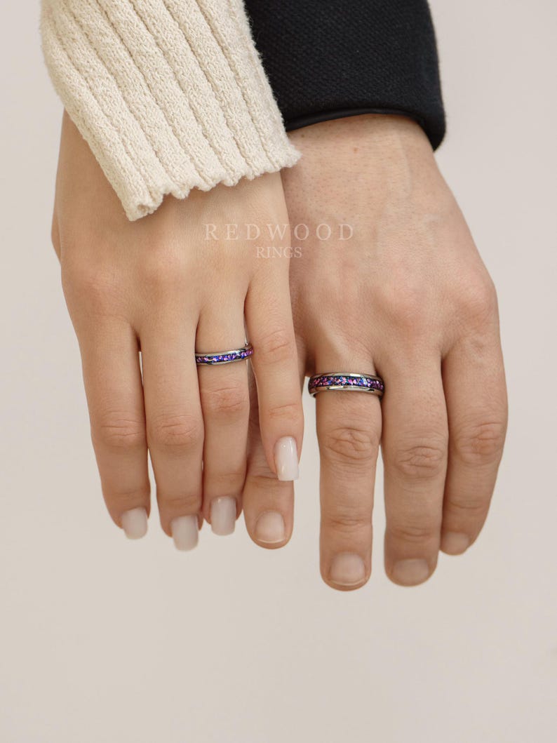 May include: Two matching rings with a band of small purple and blue gemstones. The rings are on the fingers of two people.