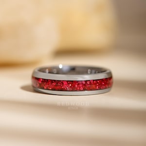 May include: A silver ring with a vibrant red inlay. The ring features a polished metal band with a central section filled with small, sparkling red fragments. The text "REDWOOD RINGS" is visible at the bottom.