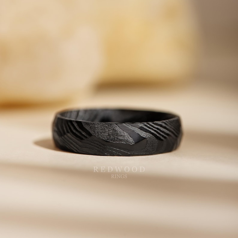 May include: A black ring with a textured surface and a unique pattern. The ring is made by Redwood Rings. The ring's design features a series of diagonal lines and a dark, matte finish, creating a modern and stylish look.