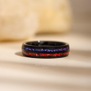 May include: A black ring with a band of red and blue inlaid stones. The ring has a polished finish and the text "REDWOOD RINGS" is visible at the bottom.
