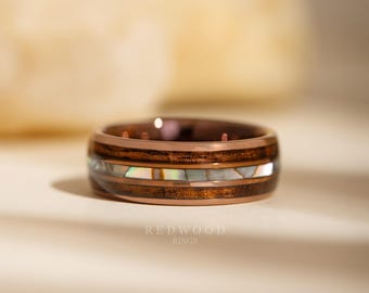 May include: A rose gold ring with a wood inlay and iridescent shell accents. The ring features a dark wood band with a lighter shell-like material in the center. The brand name "REDWOOD RINGS" is printed at the bottom.