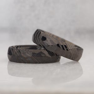 May include: Two textured, dark gray rings with a geometric pattern. One ring is resting on top of the other, creating a layered effect. The rings have a unique, carved appearance, and are displayed on a reflective surface.