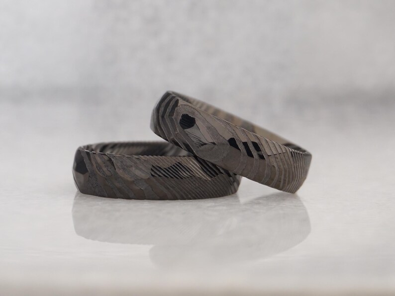 May include: Two textured, dark gray rings with a geometric pattern. One ring is resting on top of the other, creating a layered effect. The rings have a unique, carved appearance, and are displayed on a reflective surface.
