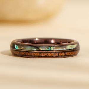 May include: A close-up of a ring with a dark brown band. The ring features a band of abalone shell and a band of wood. The ring is a unique piece of jewelry.
