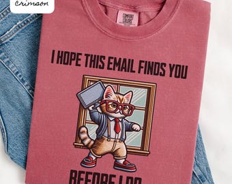 Pode incluir: T-shirt carmesim com o texto "I HOPE THIS EMAIL FINDS YOU BEFORE I DO". O design apresenta um gato de desenho animado com óculos, gravata e blazer, segurando um laptop. A t-shirt está dobrada sobre jeans azuis.