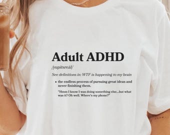 Puede incluir: Camiseta blanca con la frase "Adult ADHD" en negro, junto con una definición y frases humorísticas. El diseño incluye la frase "WTF is happening to my brain" y otros textos relacionados.