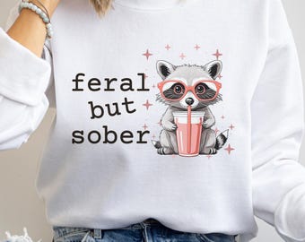 May include: White sweatshirt with the text "feral but sober" and a cartoon raccoon wearing red glasses and drinking a pink beverage. Pink stars are scattered around the raccoon.