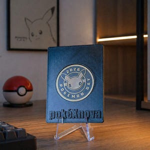 Könnte beinhalten: Ein blaues Lederportemonnaie mit einem kreisförmigen Emblem mit einem Pikachu-Design und dem Text "2026 Pokemon Day". Das Portemonnaie wird auf einem transparenten Ständer präsentiert, mit dem Wort "pokéXnova" unten geprägt.