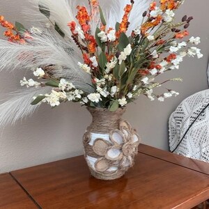 May include: A decorative vase filled with a vibrant floral arrangement. The vase is adorned with twine and a burlap flower. The bouquet features white, orange, and green flowers, along with fluffy white plumes.