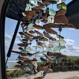 May include: A decorative wind chime featuring sea glass, driftwood, and blue beads. The sea glass is in shades of green, clear, and brown. The driftwood pieces are various sizes and shapes, creating a natural, coastal aesthetic.